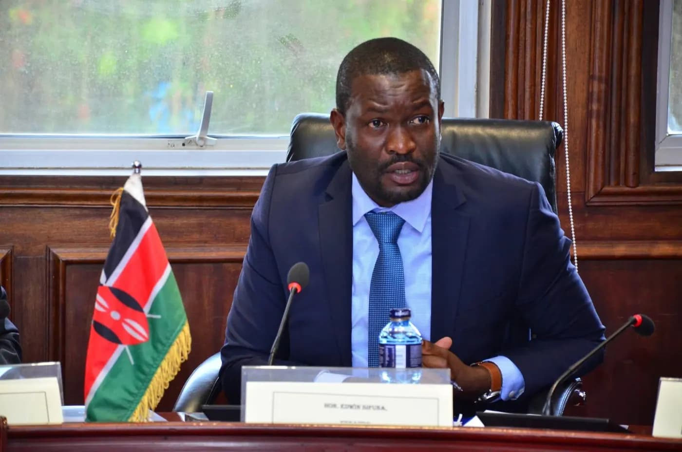 Sen. Edwin Sifuna speaking at a committee session beside a Kenyan flag.