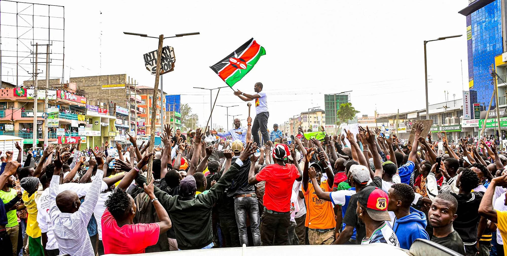 Sen. Edwin Sifuna standing on a car roof waving the Kenyan flag above a cheering crowd on a Nairobi street.